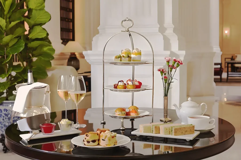 Elegant afternoon tea at Raffles Hotel Singapore with three-tier pastry stand, scones, sandwiches, and champagne on a polished table.