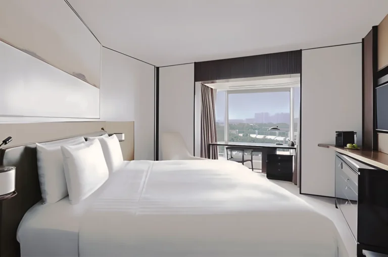 Contemporary hotel room in Singapore featuring a king‑size bed with crisp white linens, minimalist décor, large window overlooking the city skyline, and a sleek work desk with coffee machine.