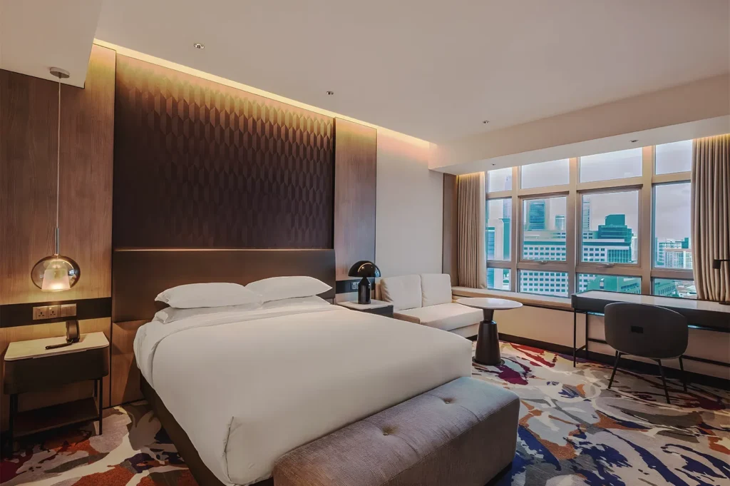 Modern luxury hotel room with a plush king bed, patterned carpet, seating area, work desk, and large windows with a city view.
