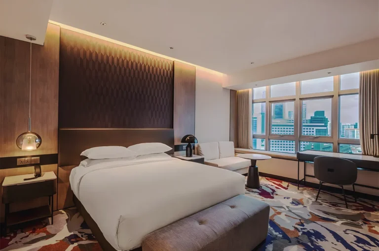 Modern luxury hotel room with a plush king bed, patterned carpet, seating area, work desk, and large windows with a city view.