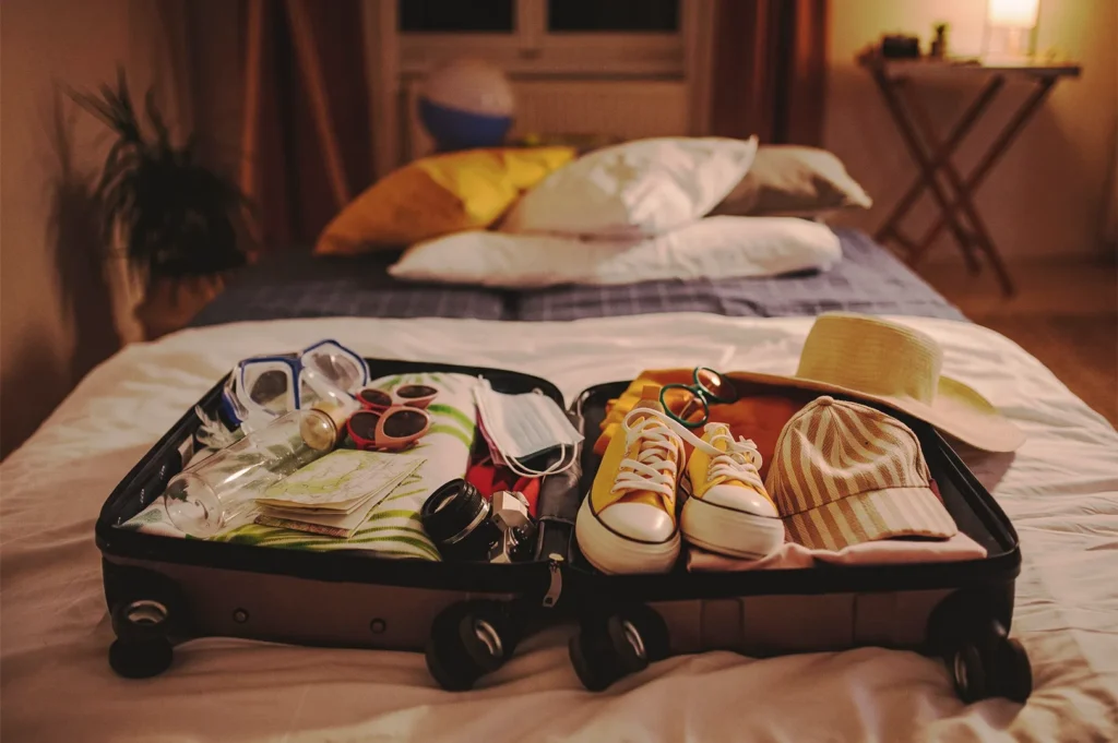 Open suitcase packed with yellow sneakers, straw hat, camera, and travel essentials on a hotel bed.