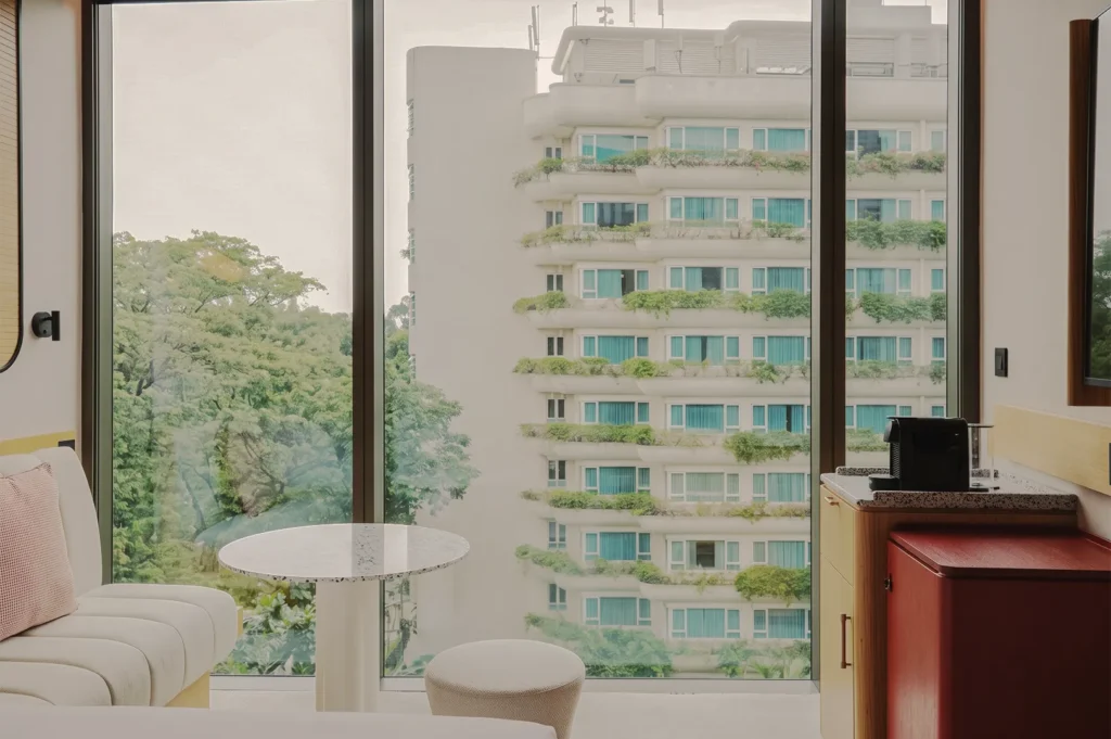 Modern hotel room interior with floor-to-ceiling windows overlooking lush green trees and a white apartment building with balcony planters.