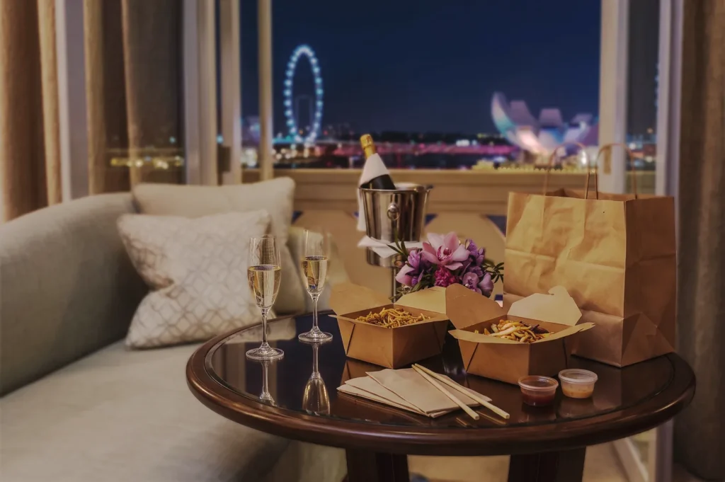 Cozy evening setup featuring champagne glasses, takeout boxes, and flowers on a table beside a sofa, overlooking the illuminated Singapore Flyer and ArtScience Museum, symbolizing upscale urban dining and relaxation.