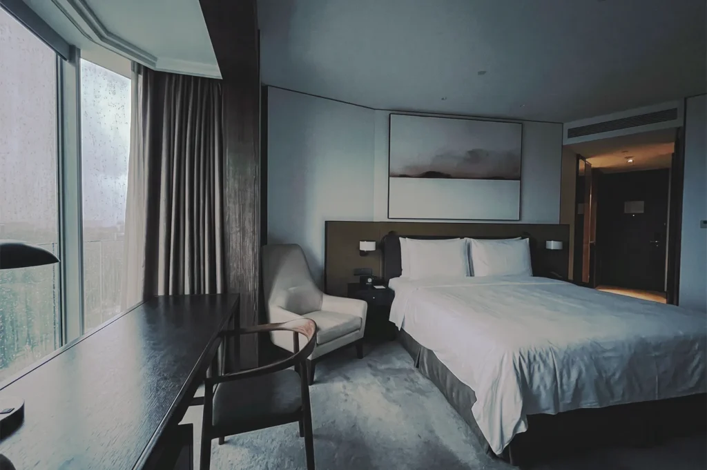 Elegant hotel room in Singapore with a comfortable bed, soft lighting, and rain‑dappled window beside a dark wooden desk and armchair, creating a serene atmosphere.