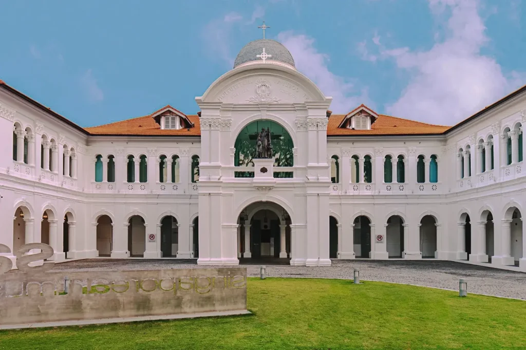 Front courtyard of Singapore Art Museum featuring colonial architecture with white arches, domed roof, and statue centerpiece under clear blue sky.