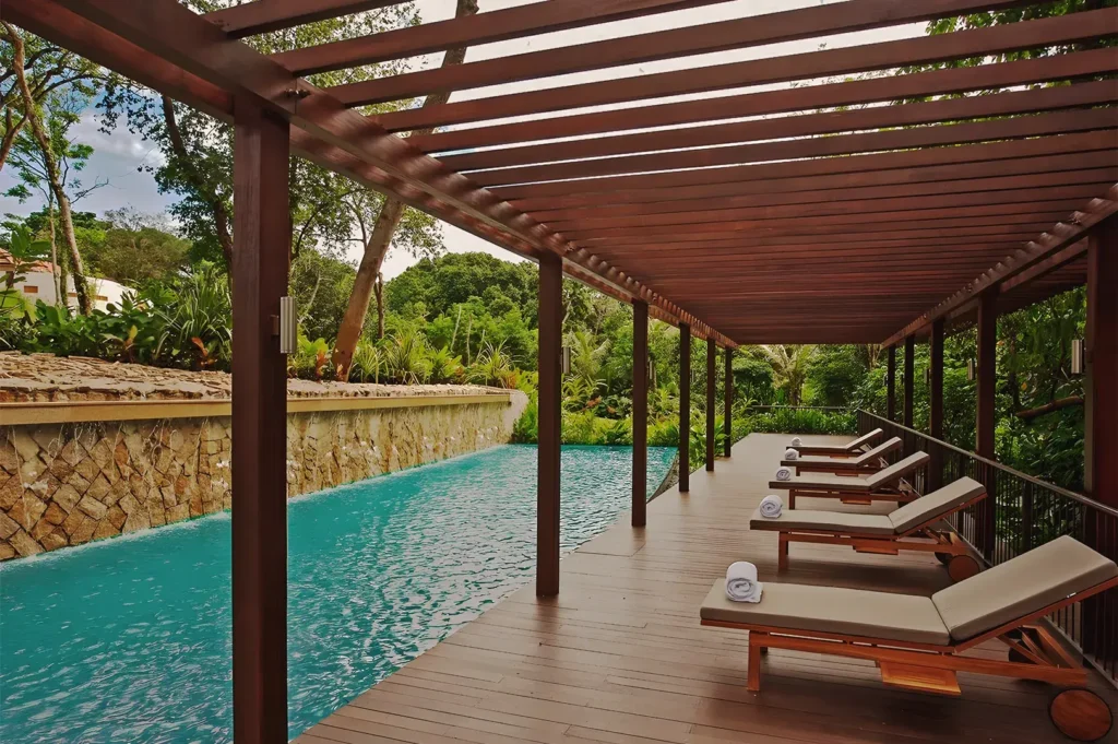 Outdoor pool deck with wooden pergola, cushioned lounge chairs, and stone wall water feature surrounded by lush greenery — peaceful resort retreat in Singapore.