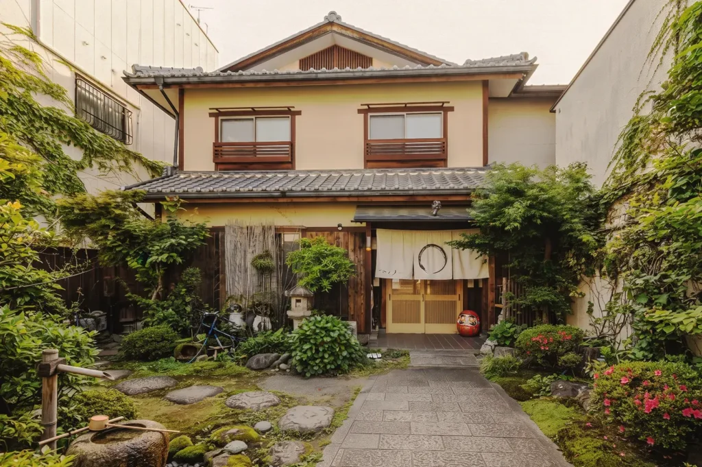 Entrance of a Kyoto‑style ryokan with noren curtain, moss garden, bamboo water feature, and Daruma doll, surrounded by lush plants and stone path.