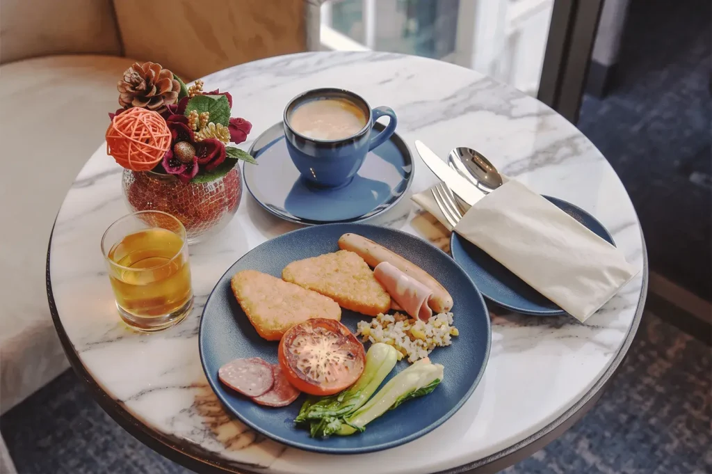 Elegant breakfast spread at Novotel Living Singapore featuring hash browns, sausage, ham, vegetables, coffee, and juice on a marble table.