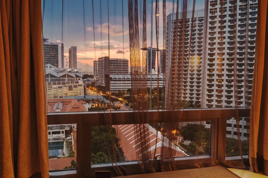 Scenic sunset view of Singapore’s skyline from a hotel window, with Marina Bay Sands visible in the distance and warm city lights glowing through sheer curtains — capturing urban elegance and evening tranquility.