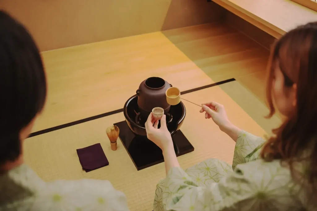Two people performing a Japanese tea ceremony on tatami mats, using traditional tea utensils and wearing yukata robes in a serene setting.