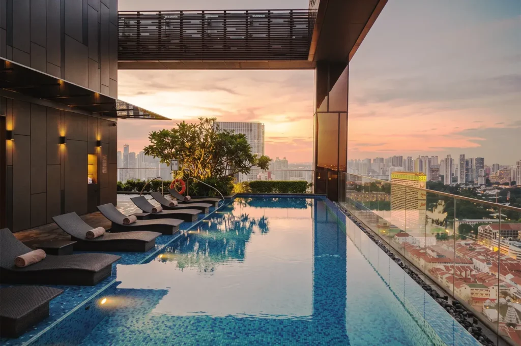 Rooftop infinity pool at sunset overlooking Singapore’s skyline, with lounge chairs and warm lighting — epitomizing relaxation, luxury, and breathtaking city views.