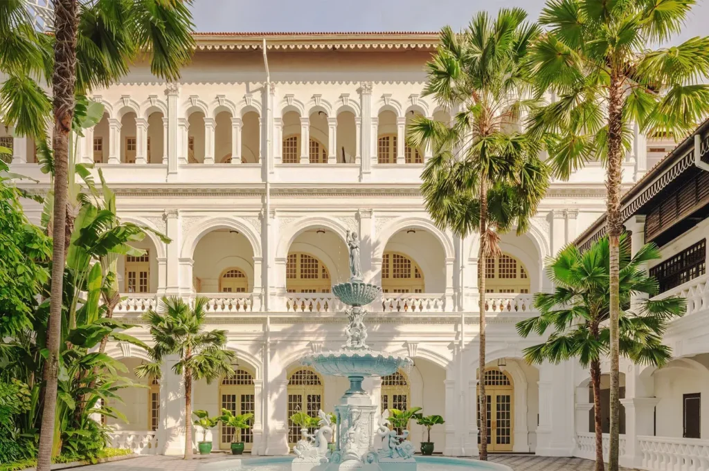 Raffles Hotel Singapore courtyard with colonial arches, palm trees, and ornate fountain surrounded by lush tropical greenery.