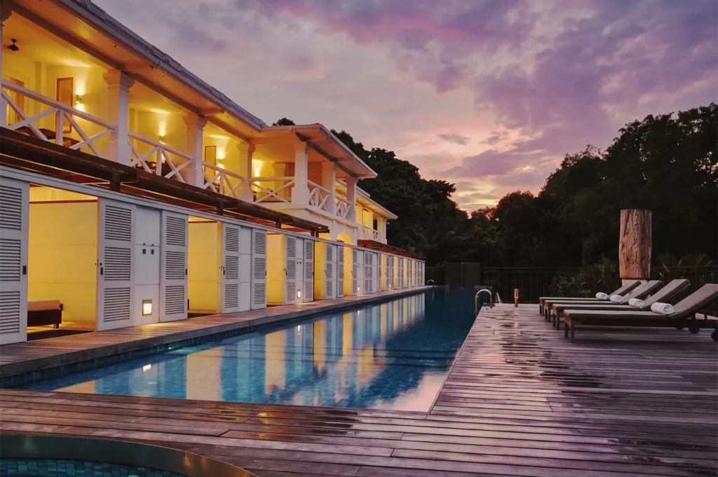 Infinity pool beside colonial‑inspired resort building illuminated at twilight, with lounge chairs and reflections of purple sunset sky — elegant boutique hotel in Singapore