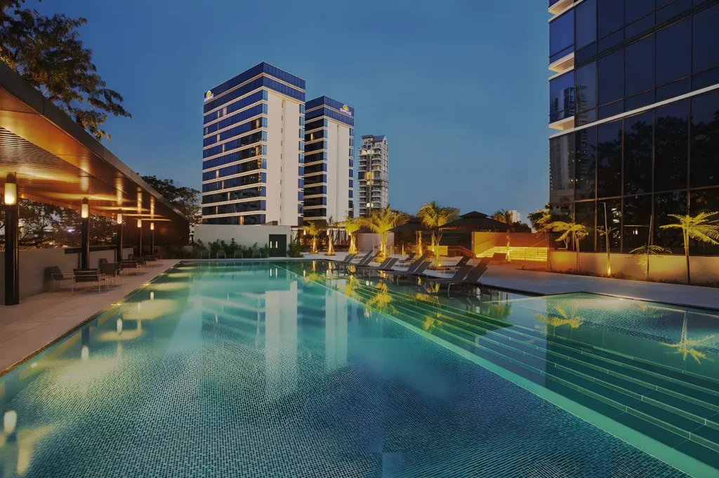 Ramada Singapore pool area illuminated at night with palm trees and lounge chairs — luxurious outdoor retreat in city hotel.