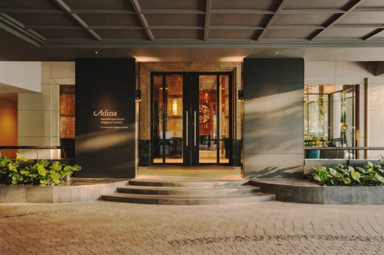 Elegant entrance of Adina Serviced Apartments Singapore Orchard featuring glass doors, modern architecture, and lush greenery under warm evening lighting.