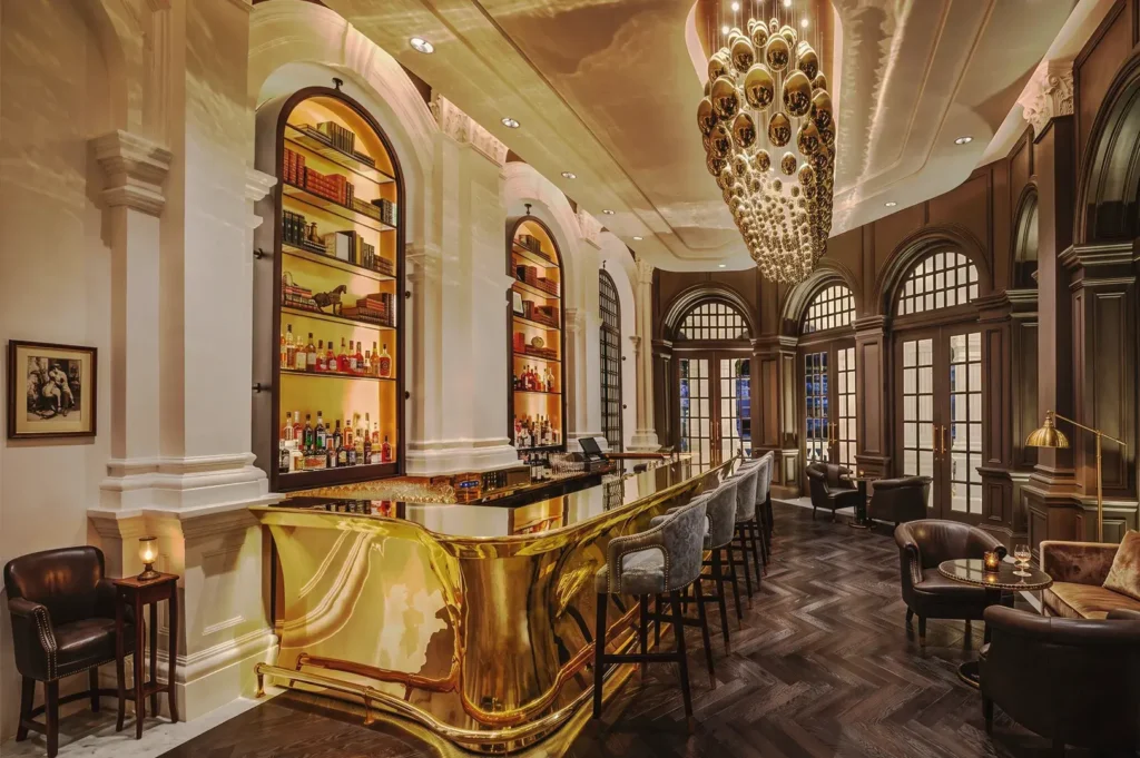 Luxurious bar interior at Raffles Hotel Singapore featuring a gleaming gold counter, arched shelves with premium liquor and books, elegant chandelier, and plush leather seating in a refined colonial setting.